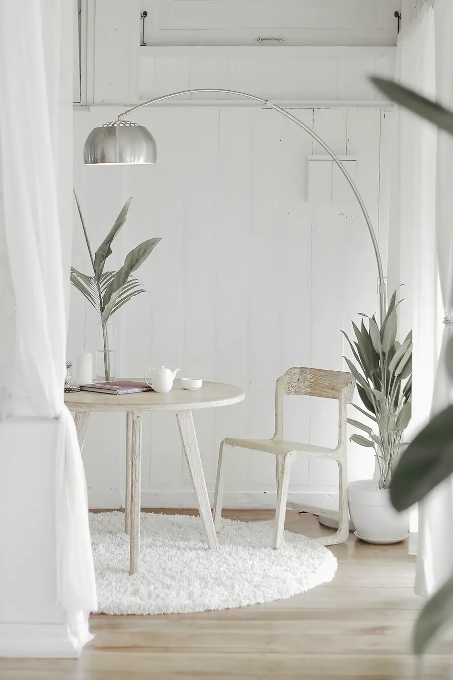 A softly lit room with sheer curtains, a small table, and leafy plants beside a chair